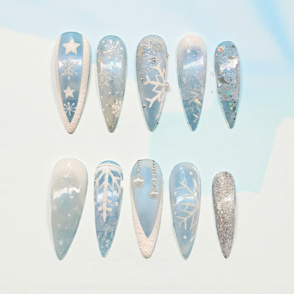 Auntie Cookies Nails: Set of 10 long stiletto press-on nails in icy blue, white, and silver with snowflake, star, and glitter designs.