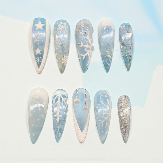 Auntie Cookies Nails: Set of 10 long stiletto press-on nails in icy blue, white, and silver with snowflake, star, and glitter designs.
