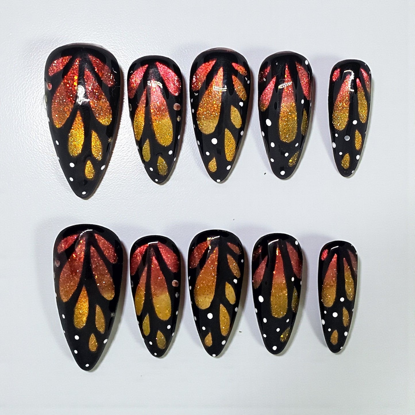 Auntie Cookies Nails almond press-on nails with black base, orange and gold glitter butterfly wing art, white dots.