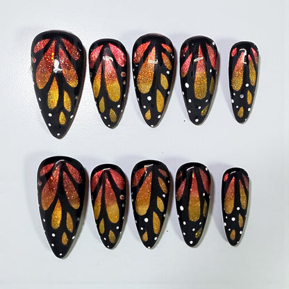 Auntie Cookies Nails almond press-on nails with black base, orange and gold glitter butterfly wing art, white dots.