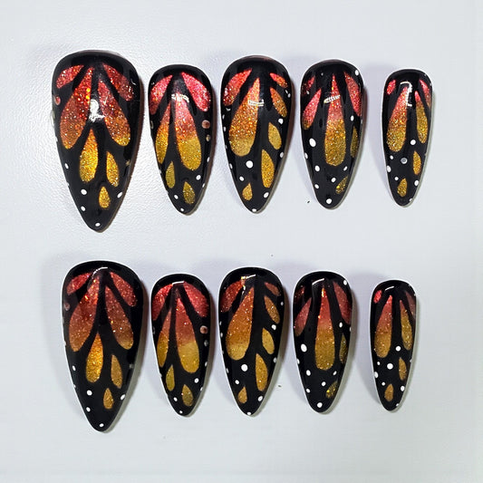 Auntie Cookies Nails almond press-on nails with black base, orange and gold glitter butterfly wing art, white dots.