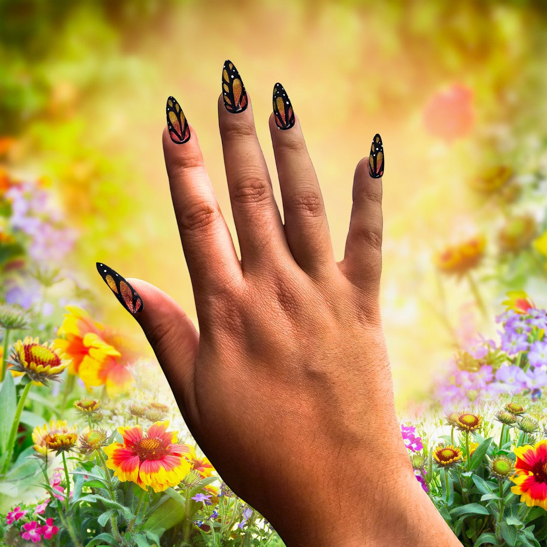 Auntie Cookies Nails almond press-on nails with black butterfly wing art, orange, red, and gold glitter in a garden.
