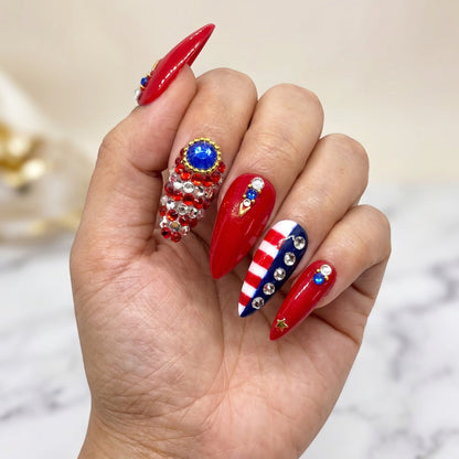 Auntie Cookies Nails stiletto press-on nails with red, white, and blue patriotic designs, rhinestones, and gold stars.