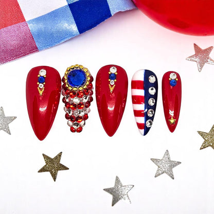 Auntie Cookies Nails stiletto press-on nails in red, white, and blue with rhinestone crystals and gold star accents.