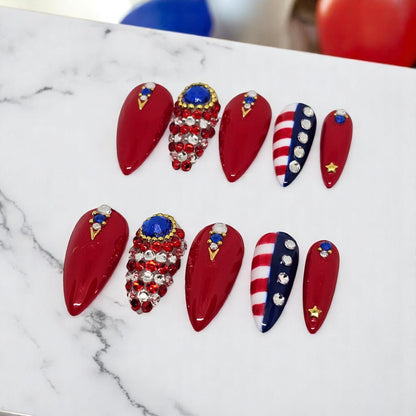 Auntie Cookies Nails stiletto press-on nails in red, white, and blue with rhinestone crystals and gold star accents.