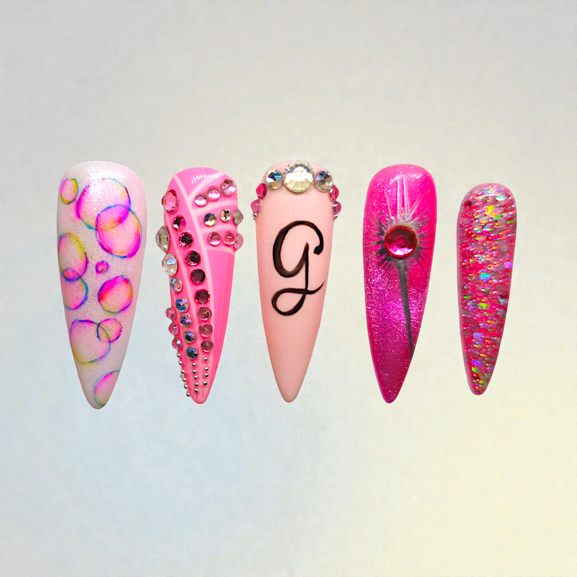 Auntie Cookies Nails: Set of 5 stiletto press-on nails in pink, white, and glitter. Designs include bubble art, rhinestones, a black 'G' initial, and a floral accent.