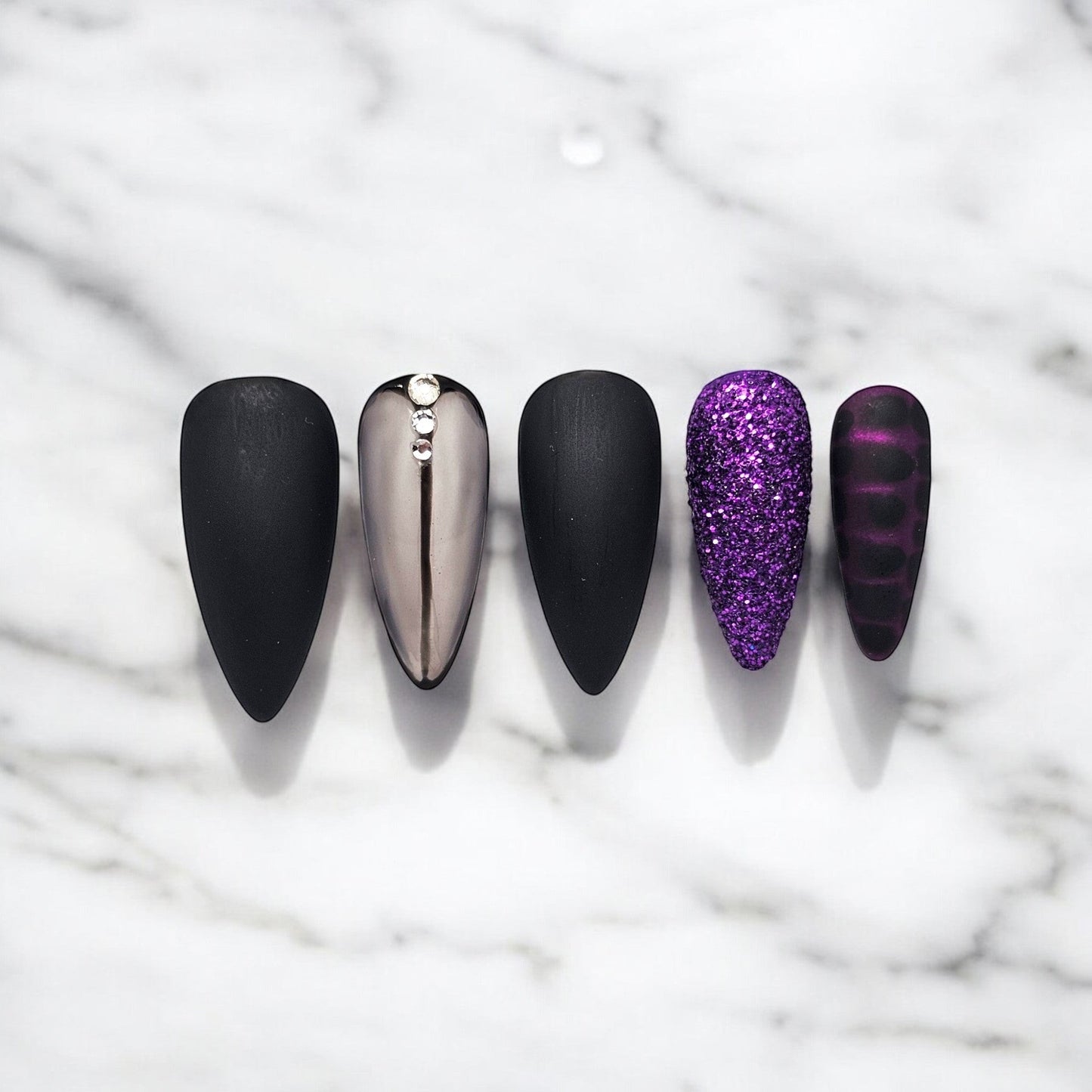Auntie Cookies Nails: Almond-shaped press-on nails in matte black, chrome with rhinestones, purple glitter, and dark purple stripes.